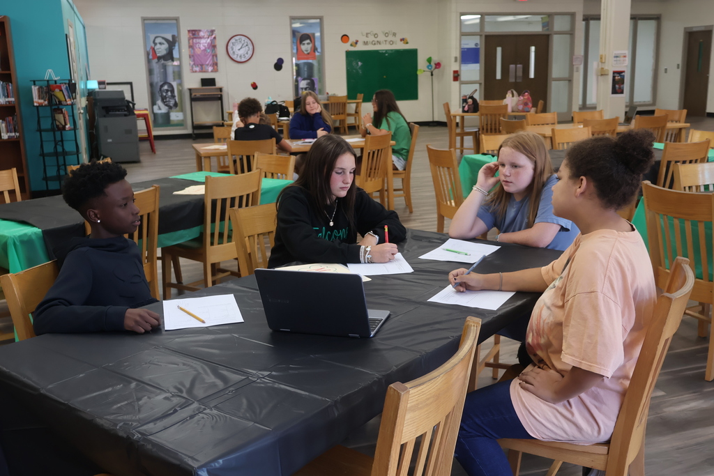 A classroom with students seated at tables. Four individuals are gathered around one table, engaging in conversation.