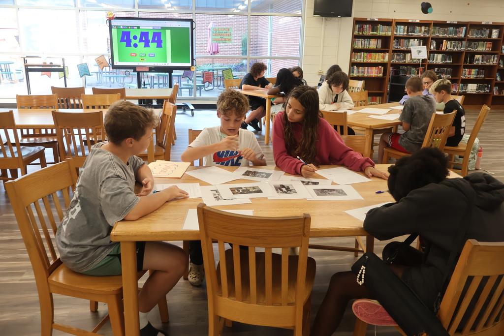 Students seated at a table in a library with papers, a monitor on the wall. Library shelves and chairs in background.
