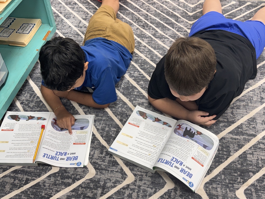 Two elementary students lying stomach-down on a grey and white patterned rug, reading open textbooks titled "Bear and Turtle Have a Race."