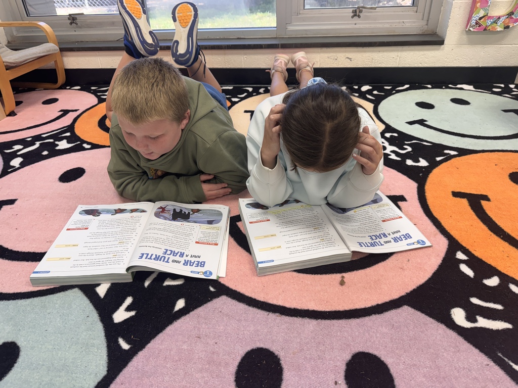 Two elementary students lying stomach-down on a colorful smiley-face patterned rug in a classroom, reading open textbooks titled "Bear and Turtle Have a Race."