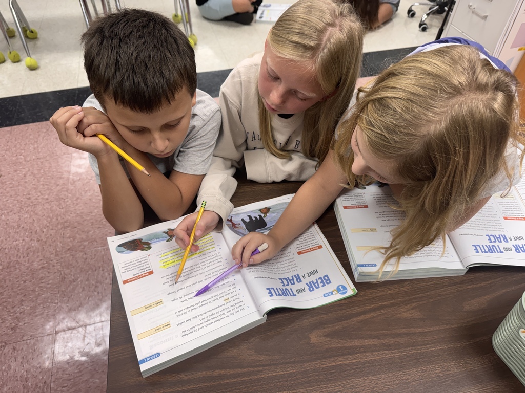Three elementary students sitting together at a desk leaning over open reading textbooks titled "Bear and Turtle Have a Race," pointing at the page with a pencil and pen as they collaborate.