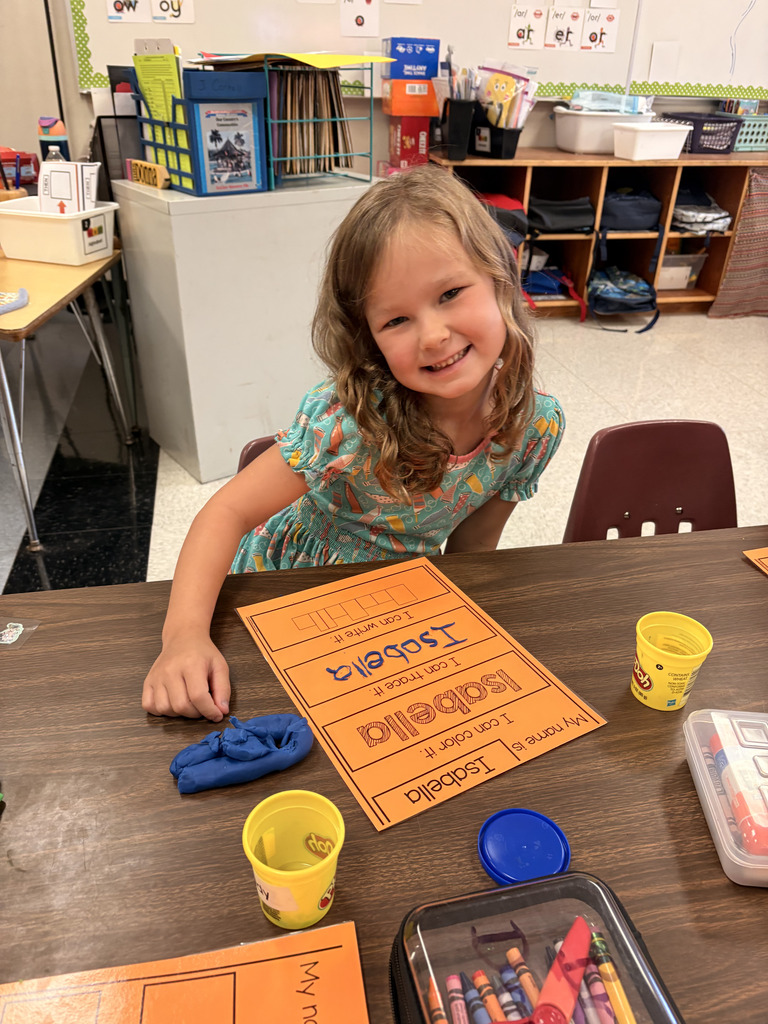A smiling young student wearing a patterned blue dress sitting at a classroom table with an orange name worksheet that reads "Isabella." Play-Doh containers and crayons rest on the table in front of her.