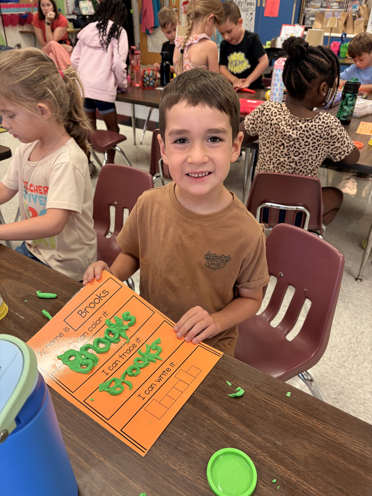 A smiling young student in a brown t-shirt sitting at a classroom table, holding an orange name worksheet that reads "Brooks." The student has shaped green Play-Doh to spell out "Brooks" on the tracing section of the page.