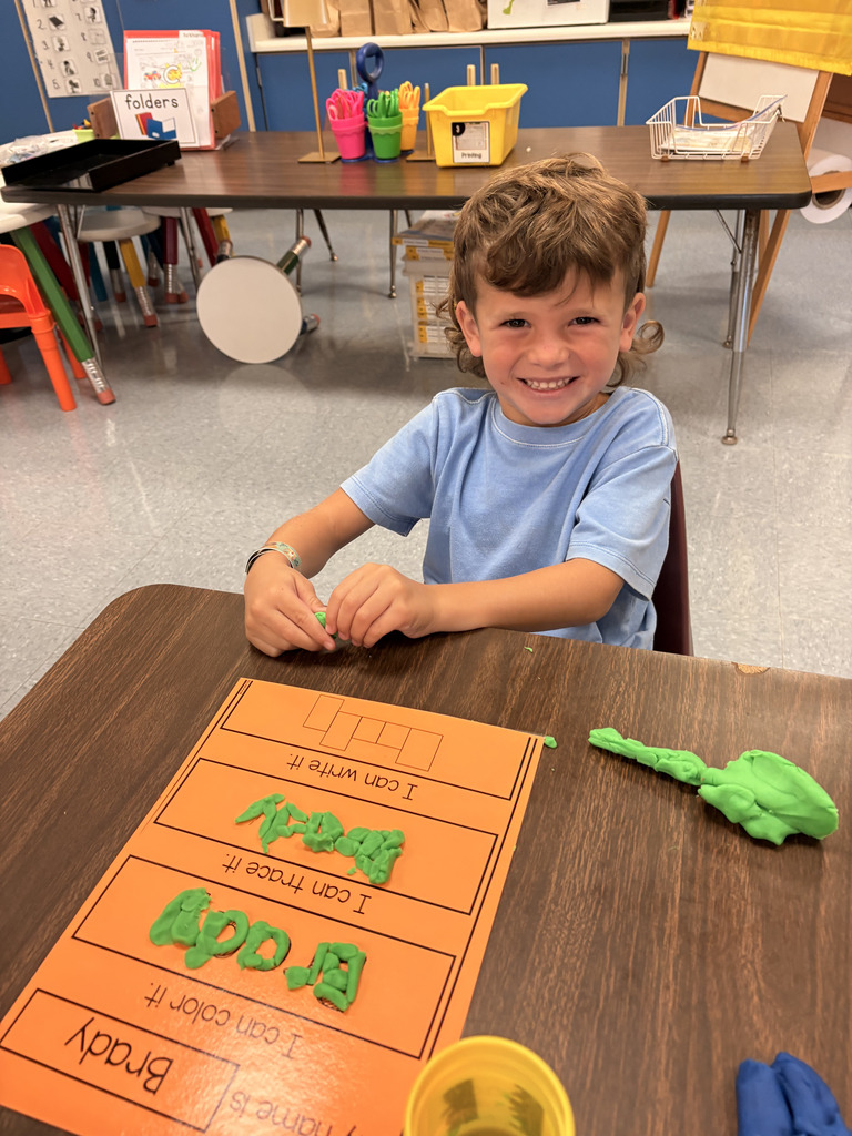 A smiling young student in a blue t-shirt sitting at a classroom desk with an orange name worksheet that reads "Brady." The student has shaped green Play-Doh into letters spelling out "Brady" on the tracing section of the paper.