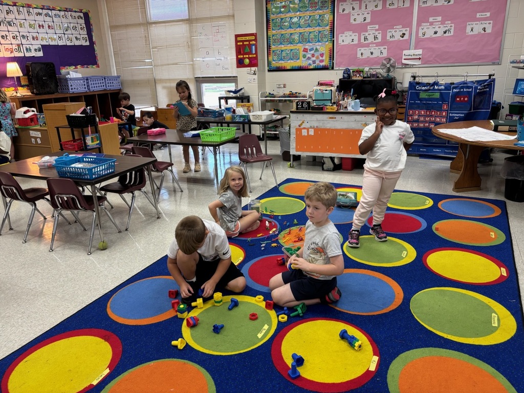 Elementary students spread out across a large, colorful circle-patterned rug in a classroom, building and playing with plastic nut-and-bolt construction toys while another student stands nearby smiling at the camera.