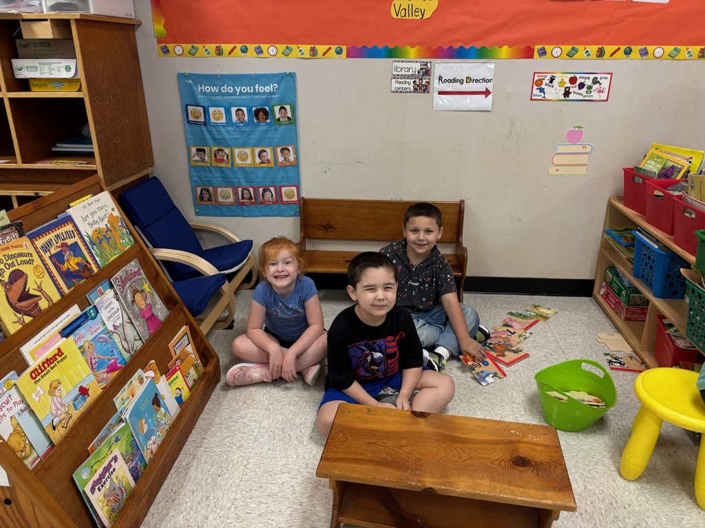 Three young elementary students sitting cross-legged on the floor in a classroom reading corner next to bookshelves display cases, a wooden bench, and educational posters.