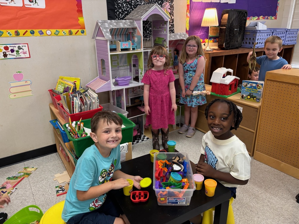 A group of young elementary students playing in a classroom play area. Two students sit at a small table with Play-Doh and cookie cutters, while two girls stand near a large wooden dollhouse in the background, and another boy peeks out from behind a shelving unit.