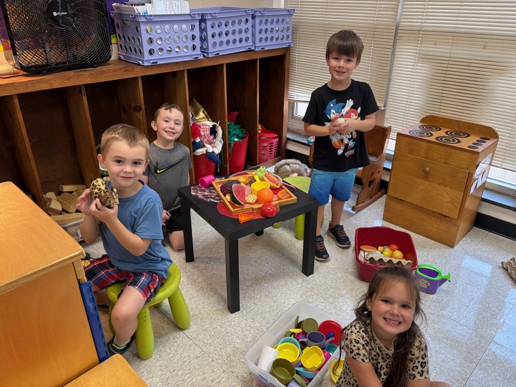 Four young elementary students smiling and playing in a classroom play kitchen area, surrounded by toy food, play dishes, a wooden stove, and small green stools.