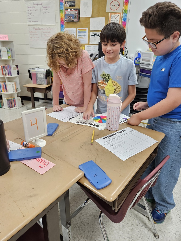 Three elementary students standing around classroom desks labeled with a station number 4 sign, reviewing tracking sheets, calculators, and a colorful spinner during a group math activity.