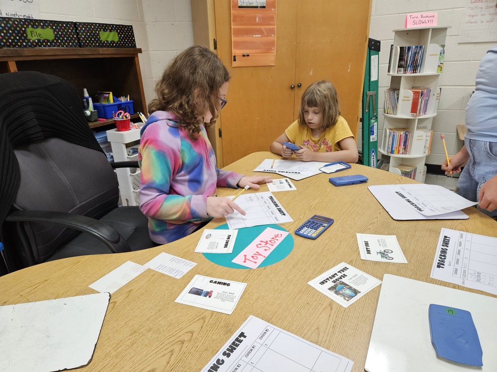 Two elementary students sitting at a semicircular table in a classroom, completing math tracking sheets with pencils using calculators and activity cards labeled Toy Store, Gaming, and Instant Tree House.