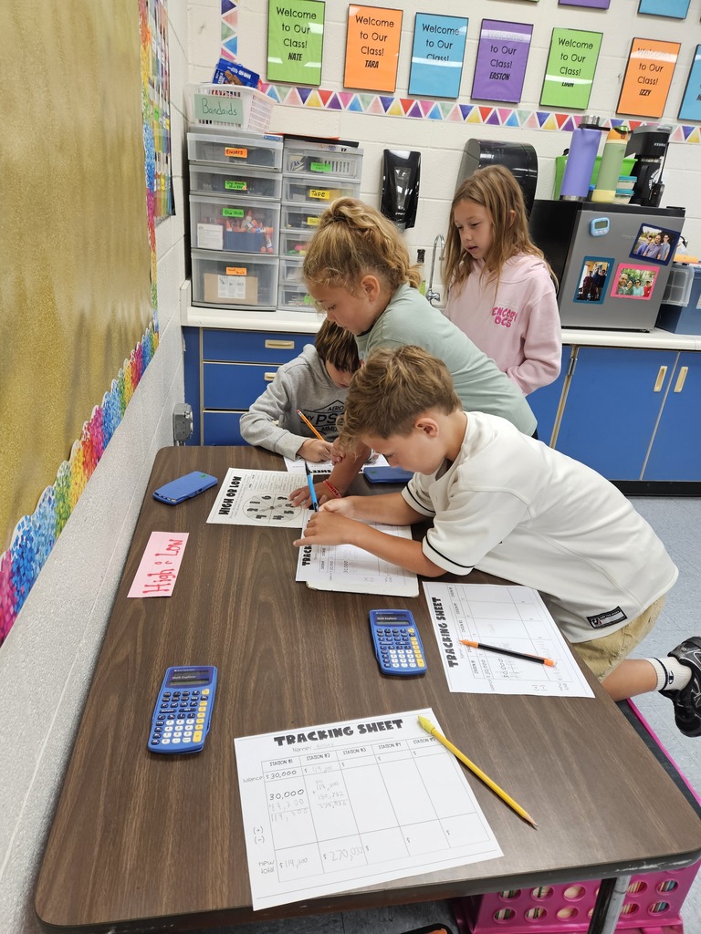Four elementary students standing and leaning over a brown activity table in a classroom, filling out math tracking sheets with pencils while using scientific calculators and spinners.