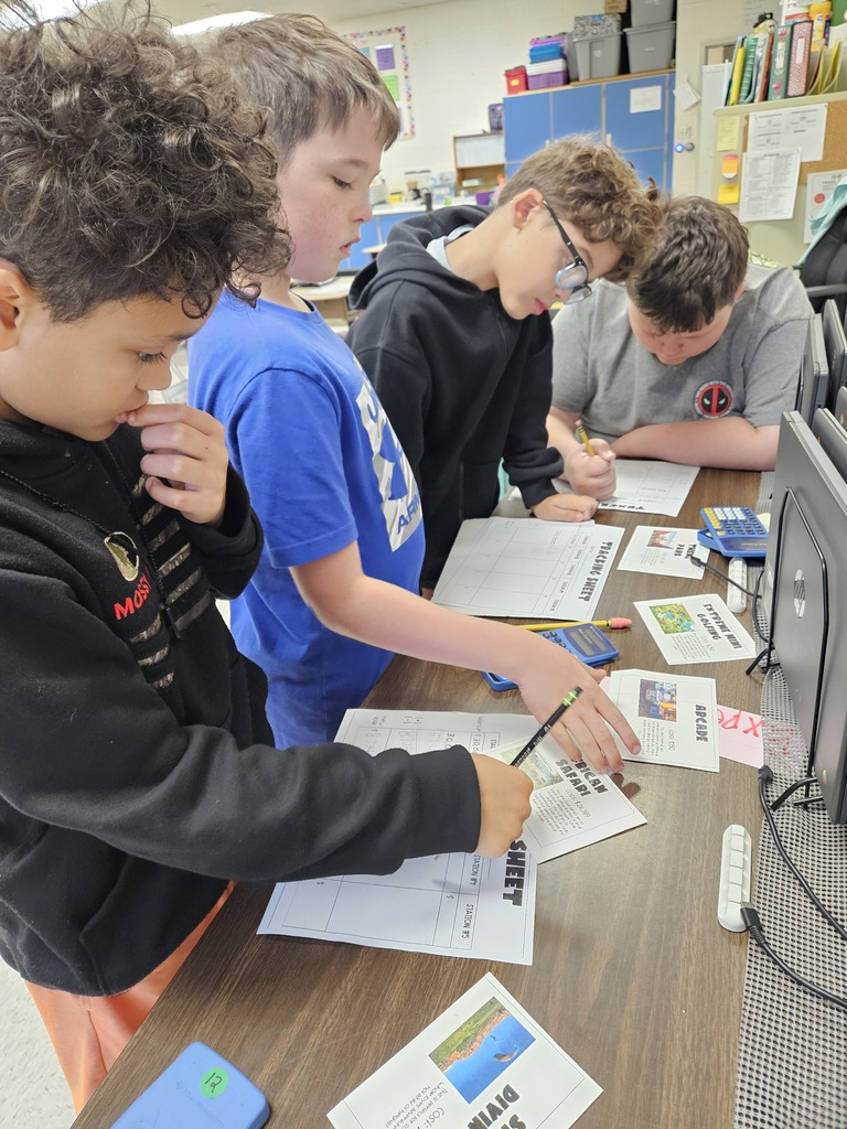 Four elementary male students standing in a row at a wooden table in a classroom, filling out tracking worksheets with pencils while working through activity cards and calculators.