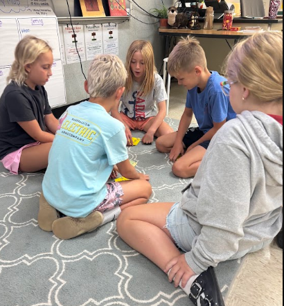 Five children seated on a patterned rug, engaged in an activity. One holds a small object.