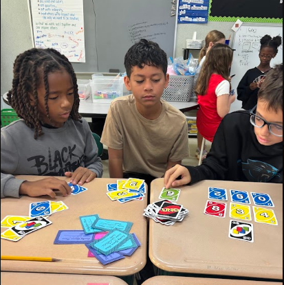 Three children sit at a table with cards and pencils. One wears glasses. Behind them, more students work at tables.