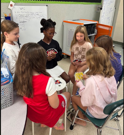 A group of children seated around a table. A girl holds a deck of cards and speaks to others.