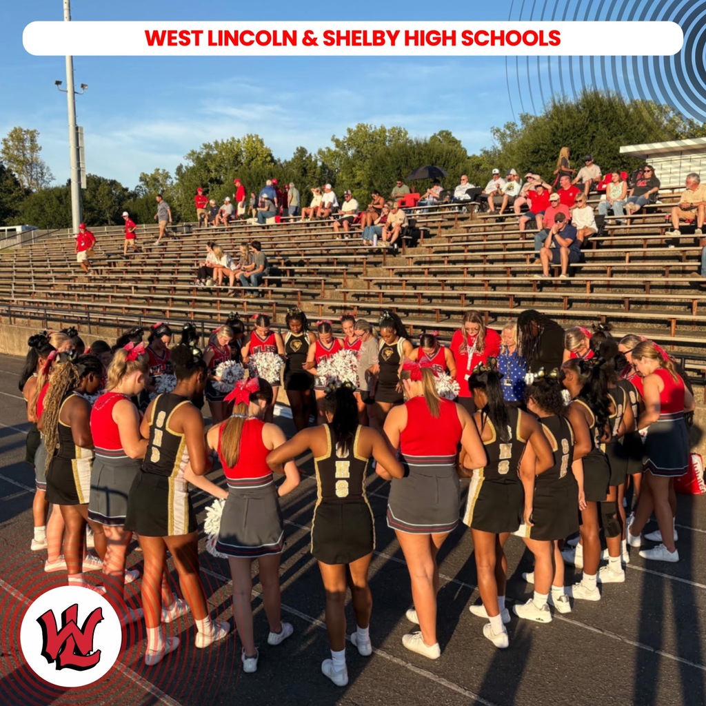 Group of cheerleaders in red and black uniforms on a field with bleachers. Background shows trees and a sky.