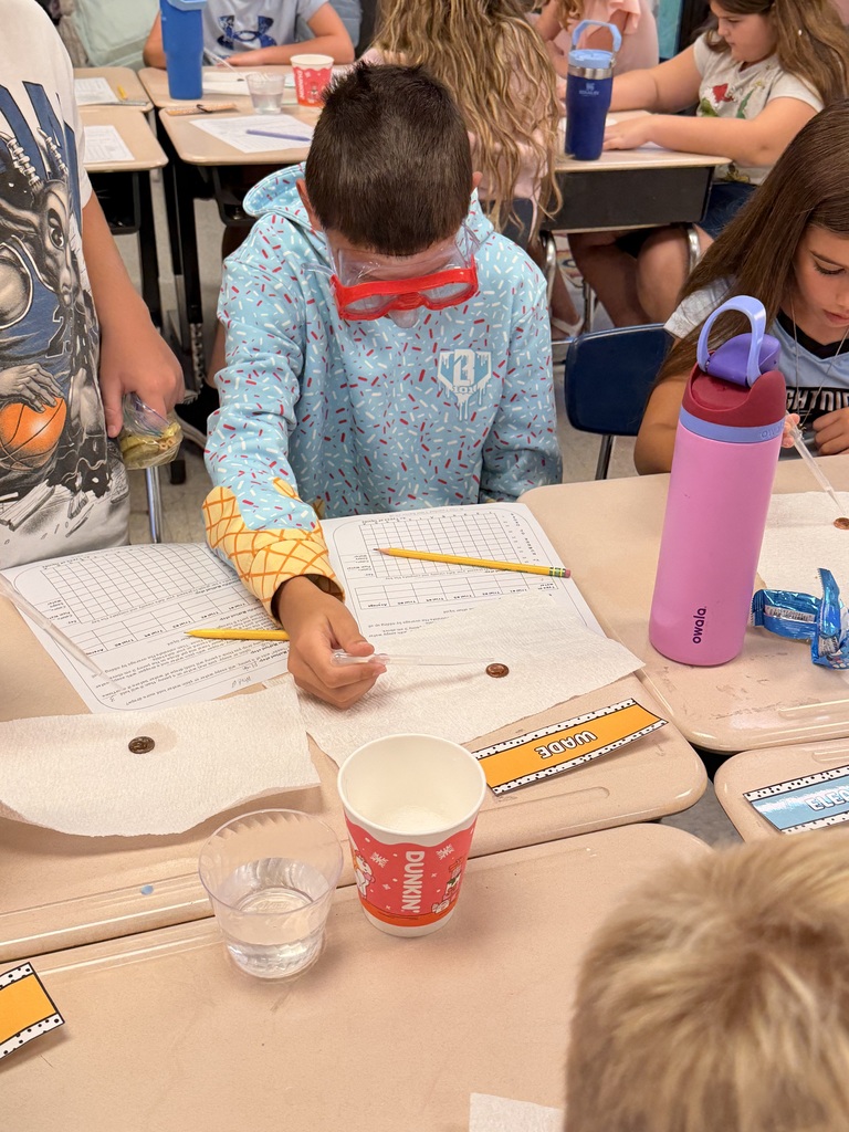 Kids seated in classroom desks, one with goggles examining something and another with a water bottle.
