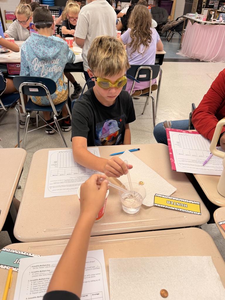 A classroom setting with students. A boy in goggles conducts an experiment. Papers, a glass, and a small container on a table.