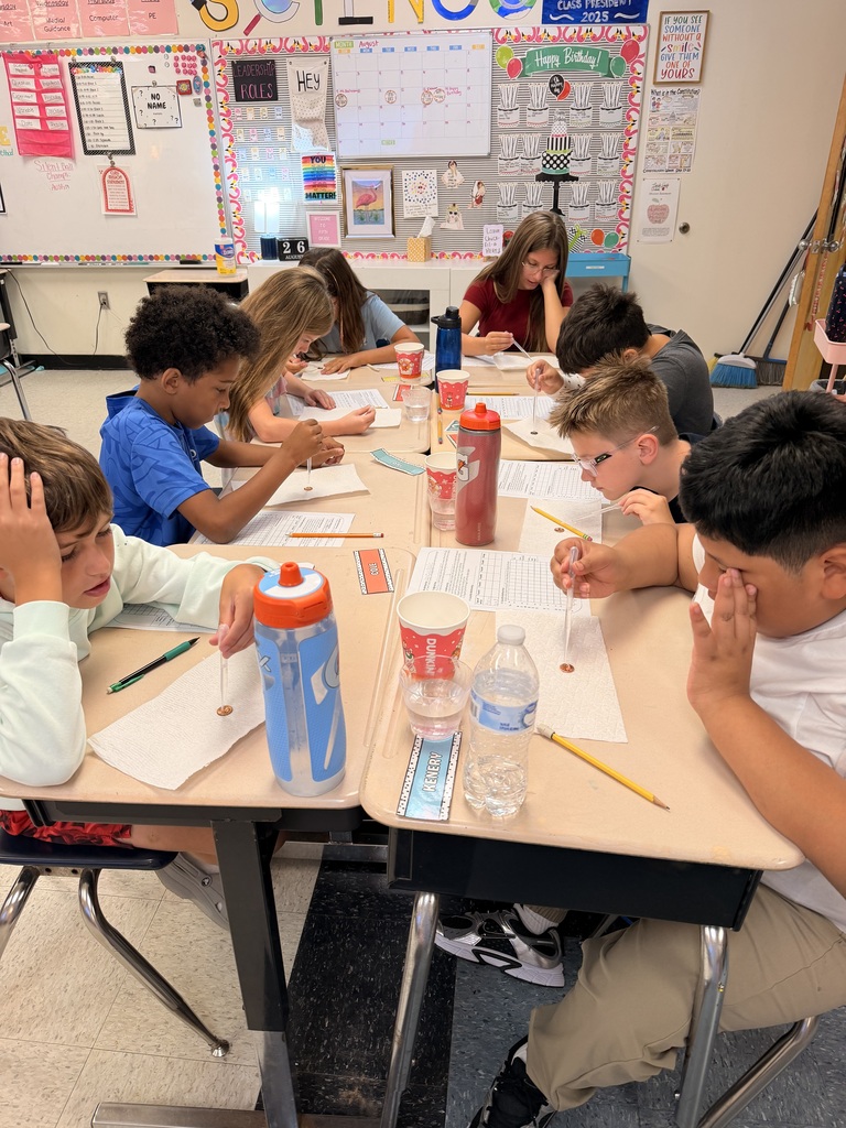 A classroom with students seated at desks, one with hands on face, others writing. Desks have bottles, pencils, paper. Whiteboard and bulletin board in background.