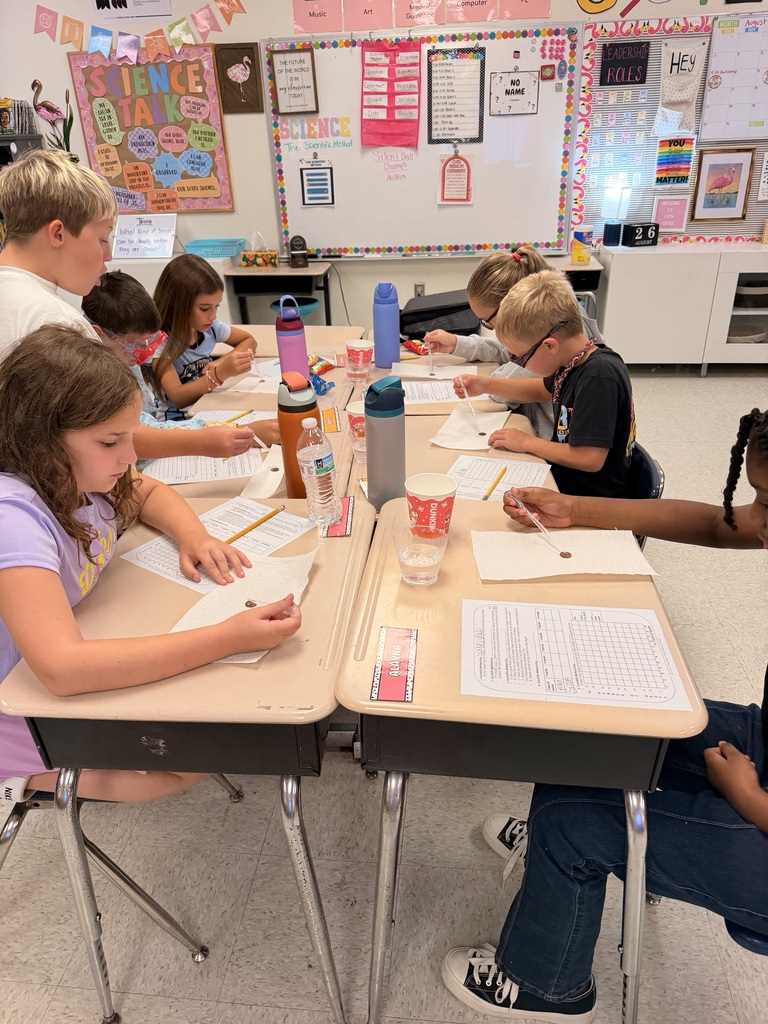 Students sit at desks in a classroom, working on papers and using pens, with various items on the desks.