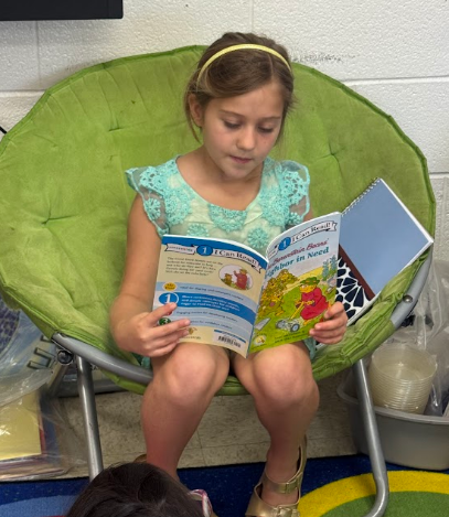 A young girl sits on a green chair reading a book. She wears a blue dress and sandals. Behind her, a wall, floor, and storage items are visible.