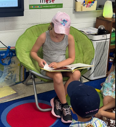 A child wearing a cap sits in a chair, reading a book. Another child sits in front, wearing a hat.