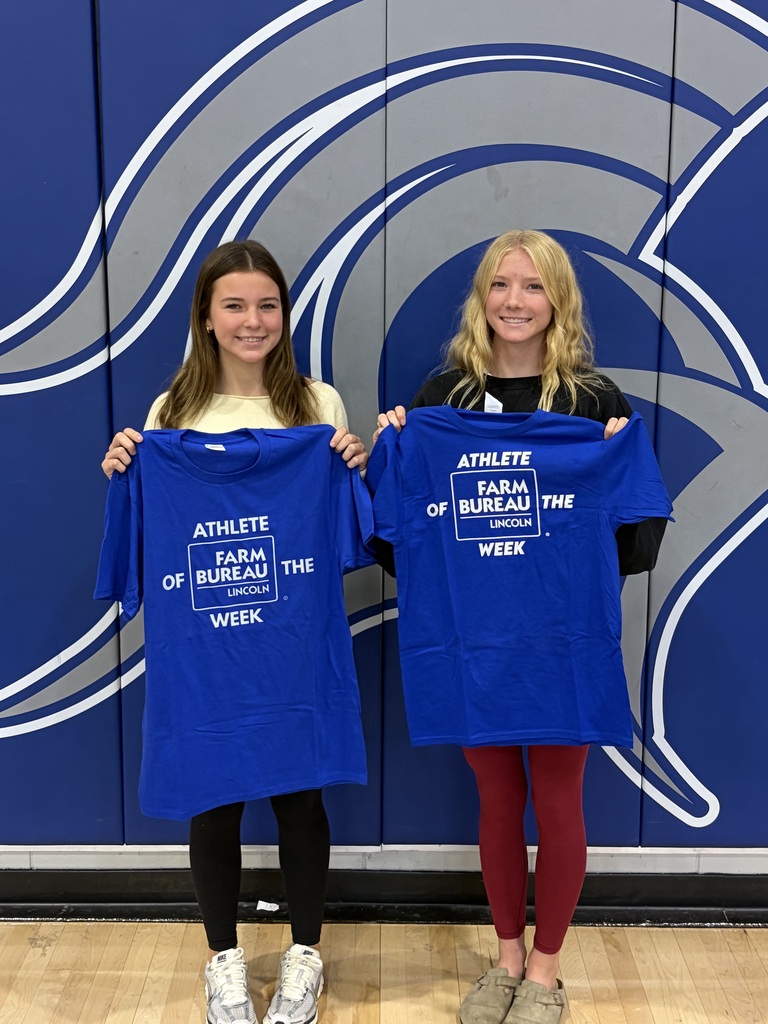 Two women in a gymnasium, holding matching blue t-shirts with white text, standing before a blue wall.
