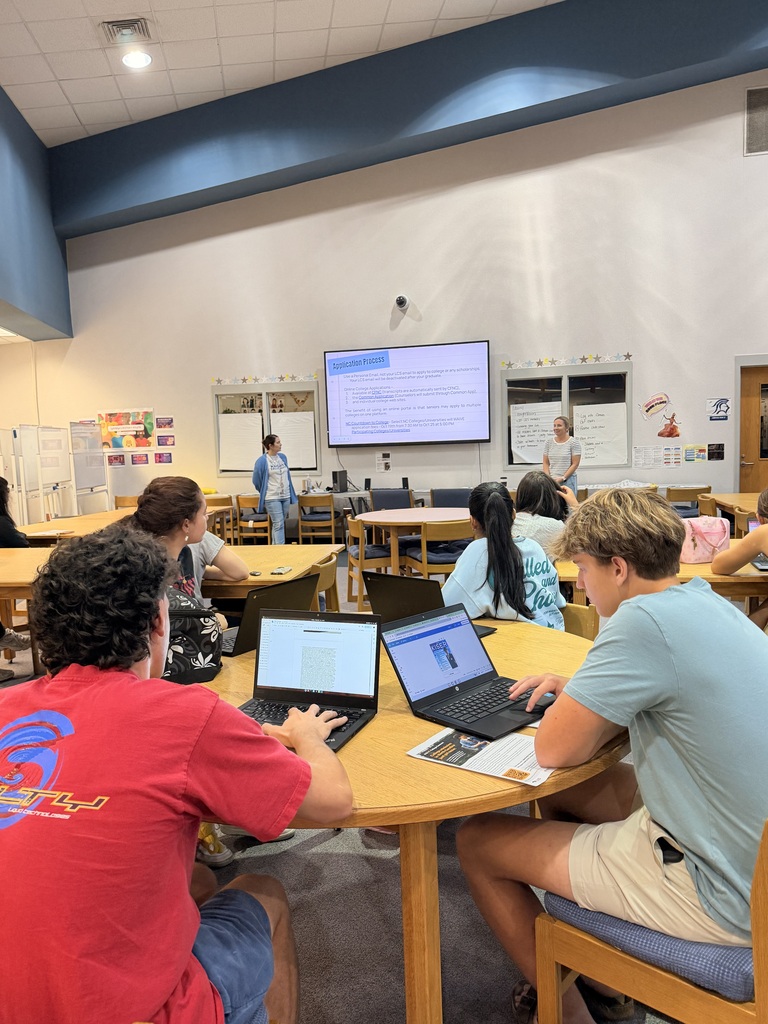 People sit at tables in a classroom, with laptops open on the tables and a screen projector behind them.