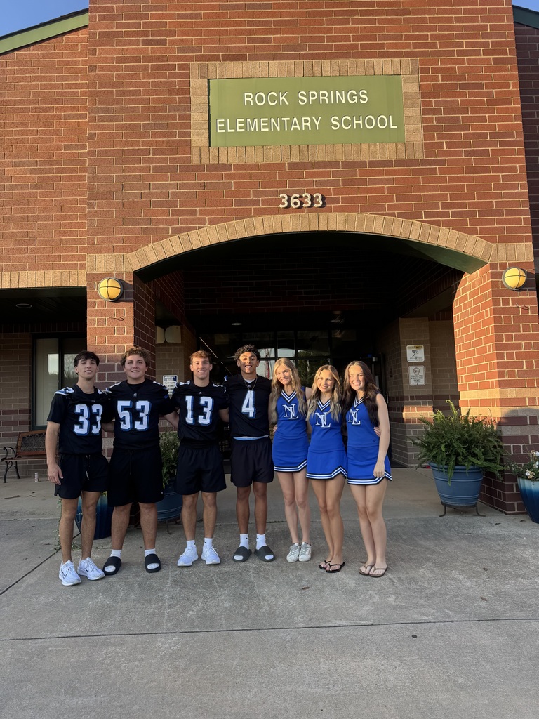 A group of students in blue uniforms stand in front of a brick building, with a sign reading "ROCK SPRINGS ELEMENTARY SCHOOL" above.