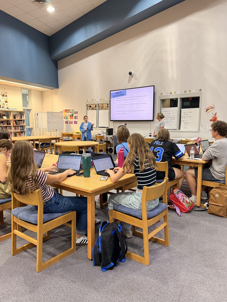 In a classroom, students use laptops at tables, a teacher stands in front of a screen.
