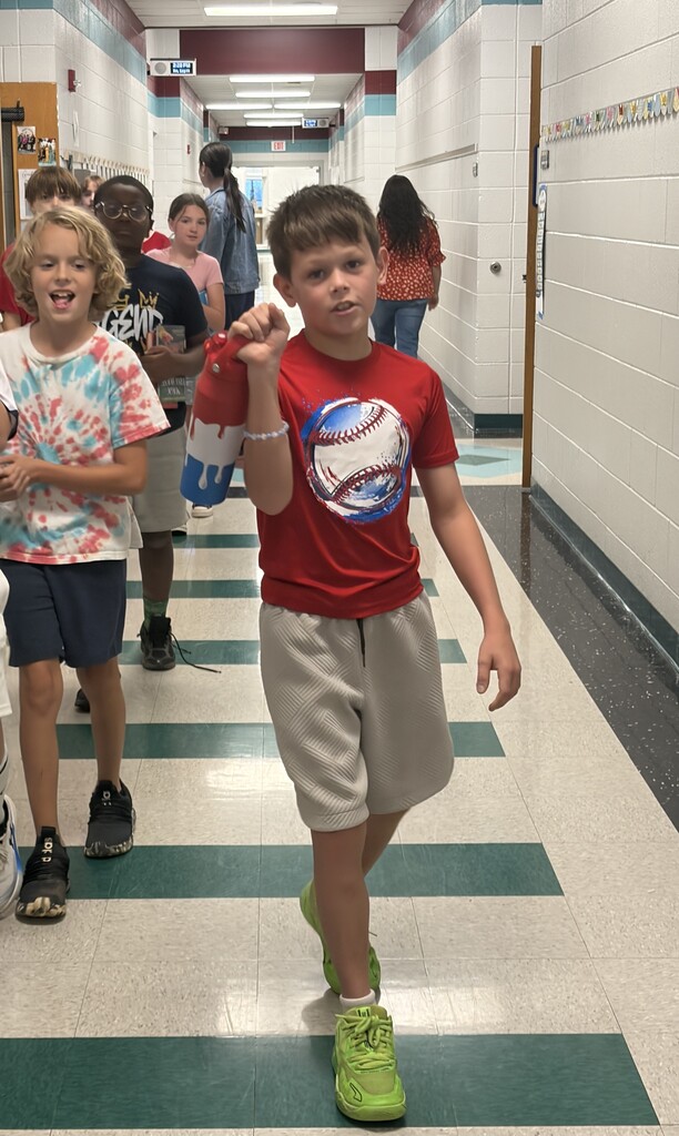 School hallway with children walking. A boy in a red shirt walks forward, making a fist. Others follow.