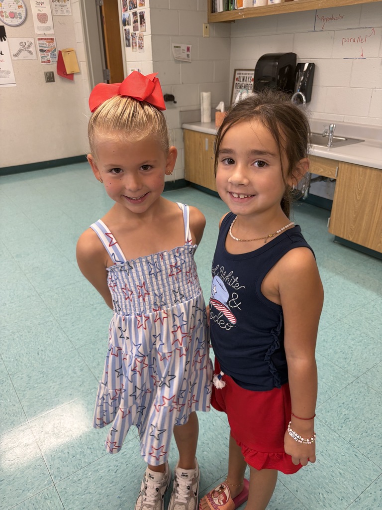 Two young girls stand in a room with a tiled floor, one with a red bow. They wear colorful outfits. A sink and cabinets are in the background.