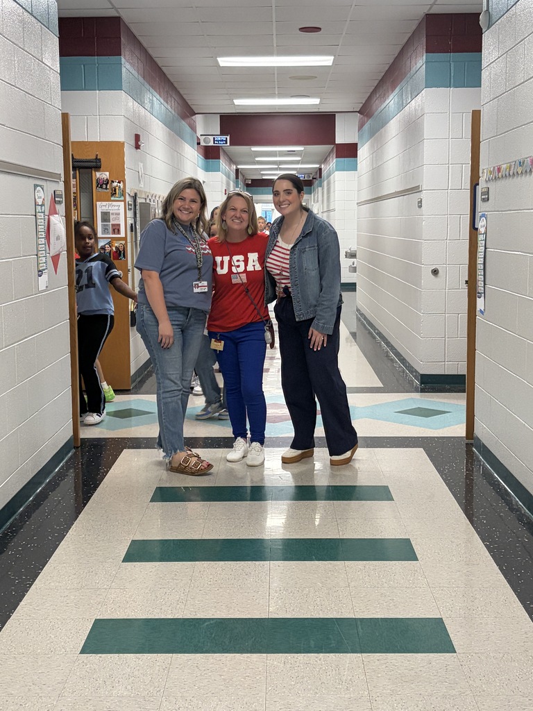 Three women stand in a hallway with white walls and green-striped floor tiles. Two women wear sneakers, one wears sandals. One woman in red shirt says "USA".