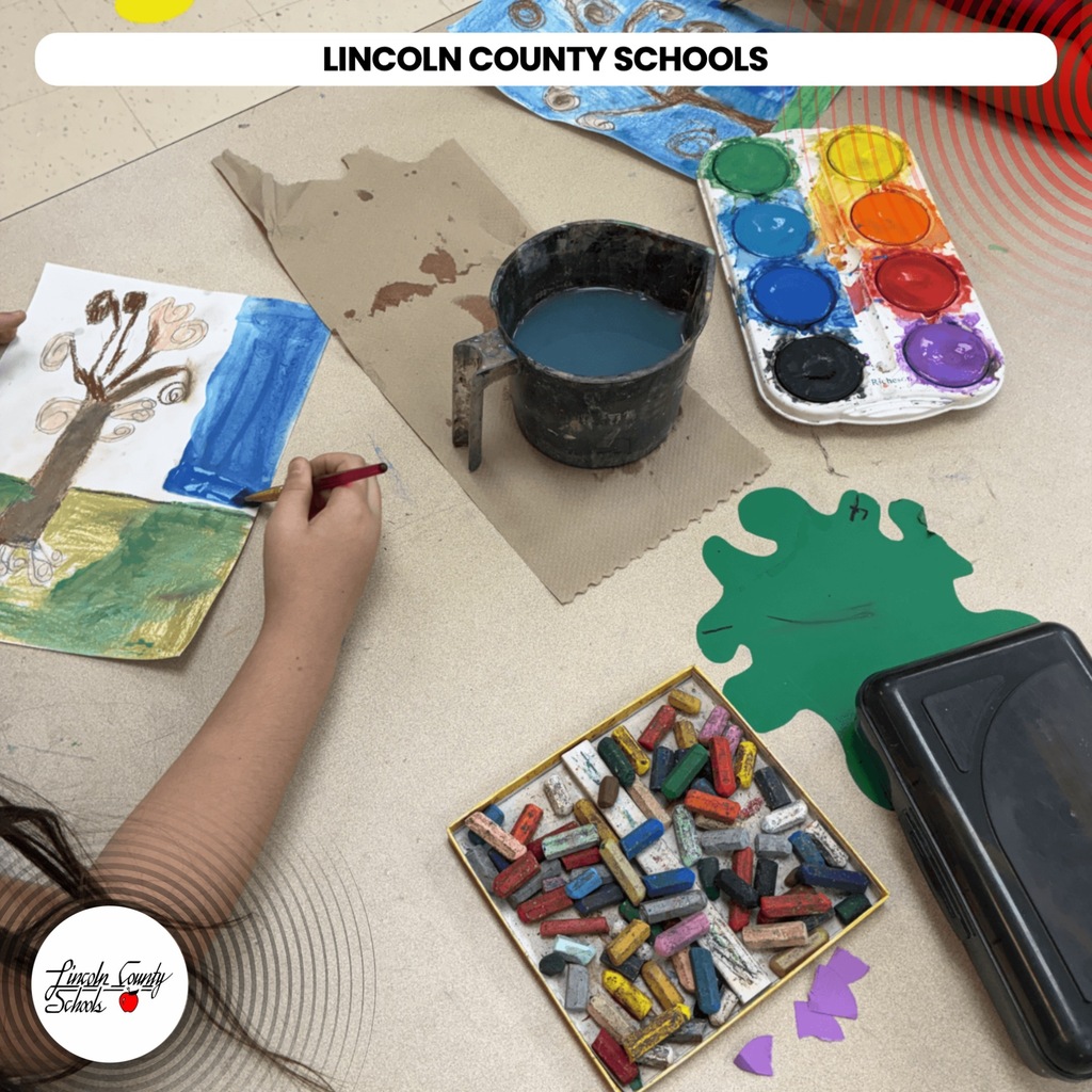 A child in an art classroom draws on a piece of paper with a red marker.
