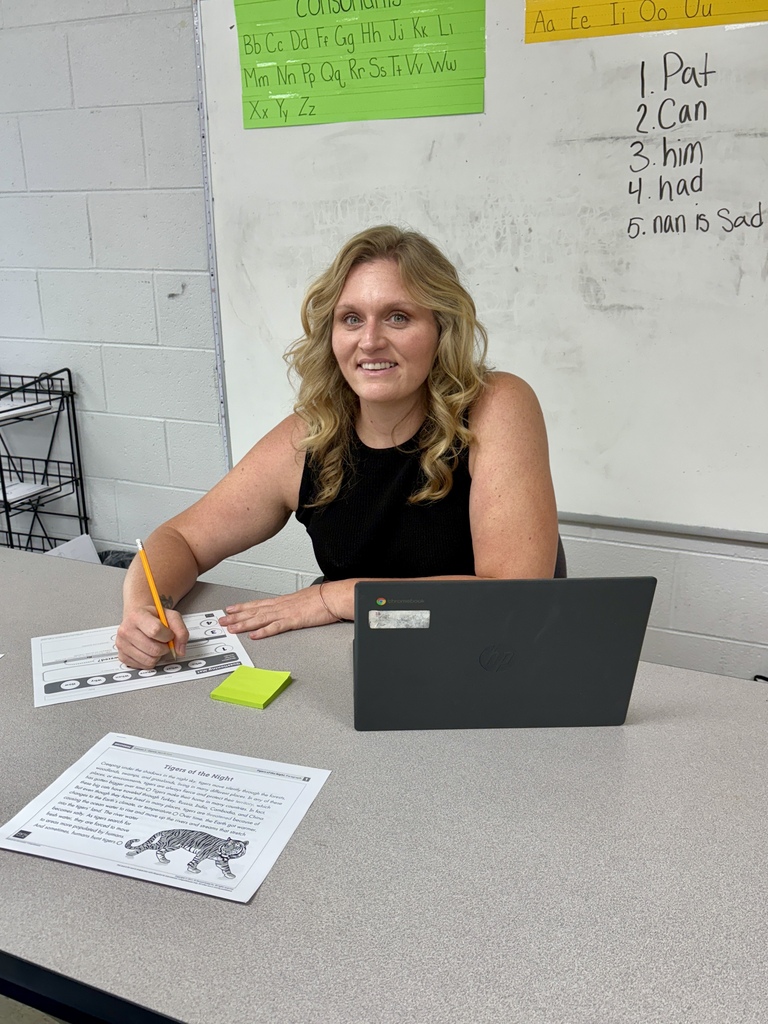 A woman with blonde hair sits at a table with a laptop, holding a pencil and smiling. Papers and a post-it are on the table. Behind her is a whiteboard.