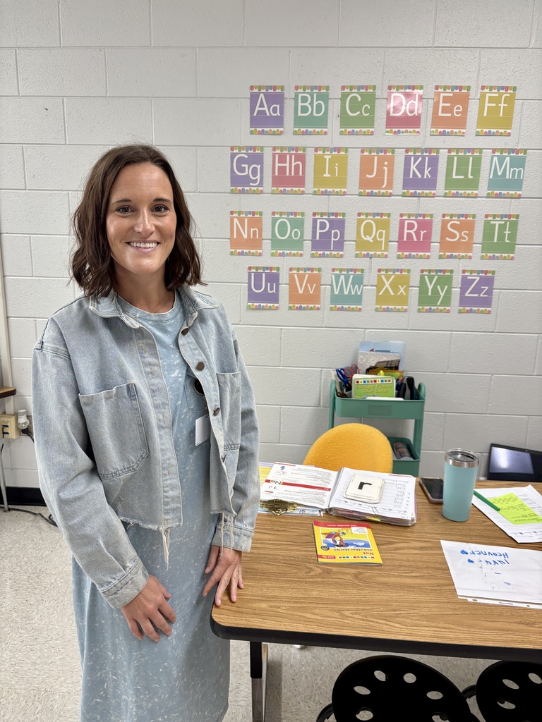 A woman stands by a table with books, papers, and a cup. Behind her, a wall has letters and numbers.