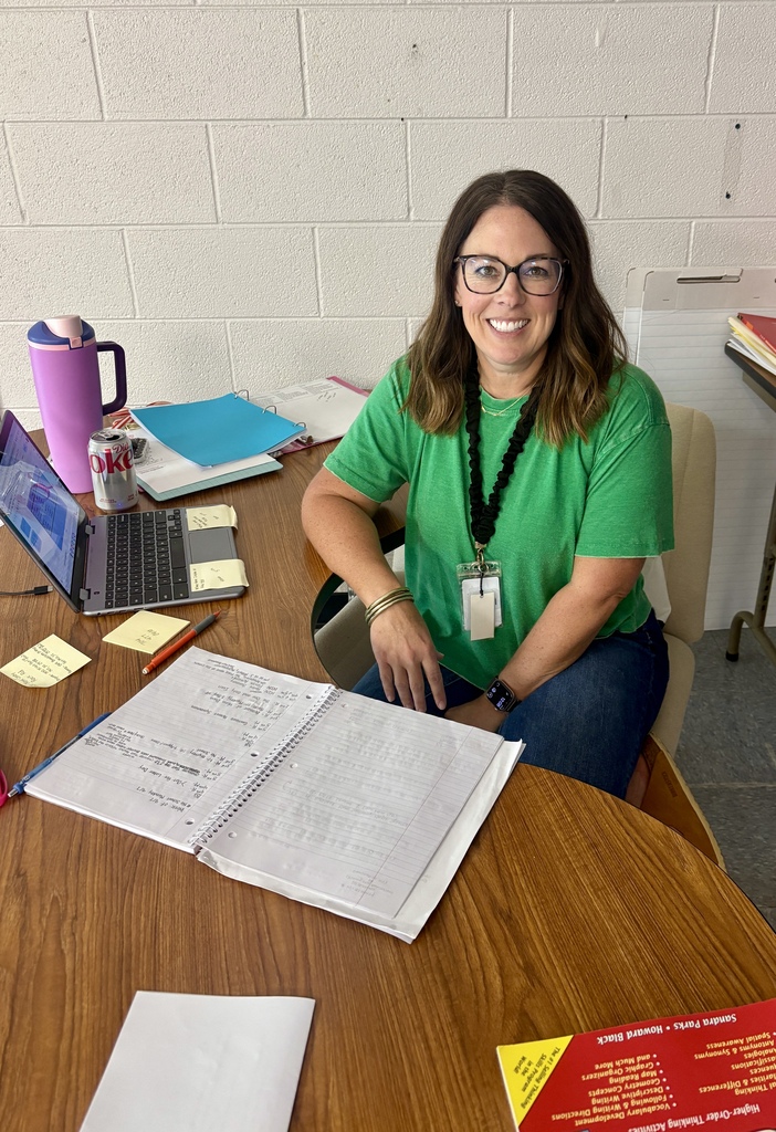 Woman sitting at a desk wearing a green shirt, smiling. Papers, pens, and a laptop are on the desk.
