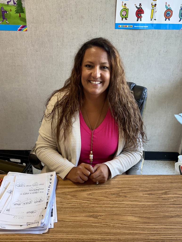 Woman in pink top with long hair sits at a desk with papers in front, smiling.