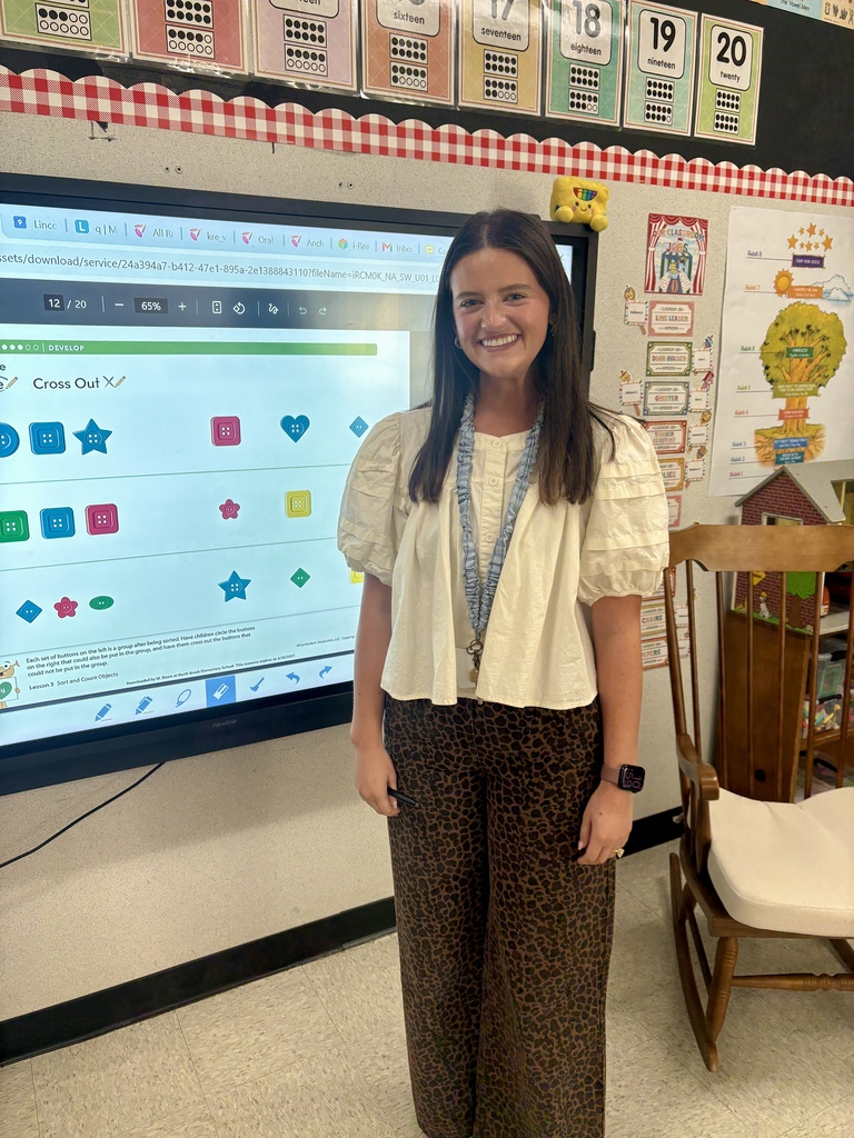 A woman stands in a classroom with a large screen behind her. The screen displays colorful icons.