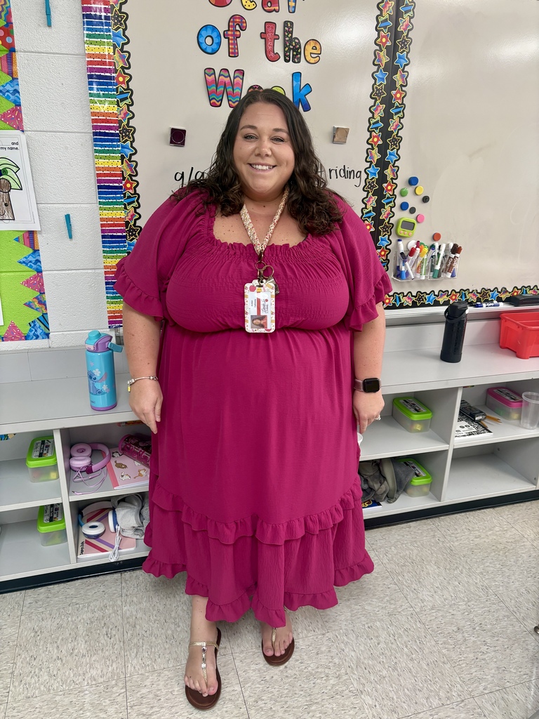 A woman in a purple dress stands in a classroom, smiling at the camera, with shelves and a bulletin board behind her.