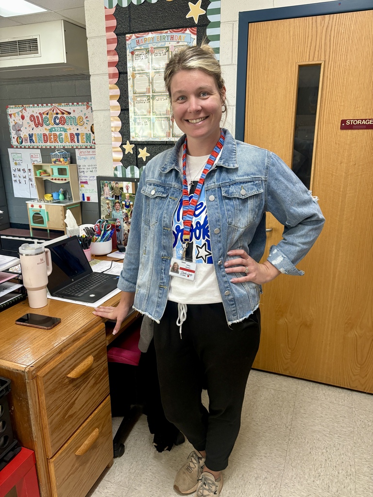 Woman with blonde hair stands in classroom, wearing a denim jacket and black pants, smiling with hands on hips.