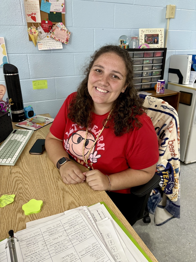 Person in red shirt with a flowery design sits at desk. The desk has a computer keyboard, phone, and papers.