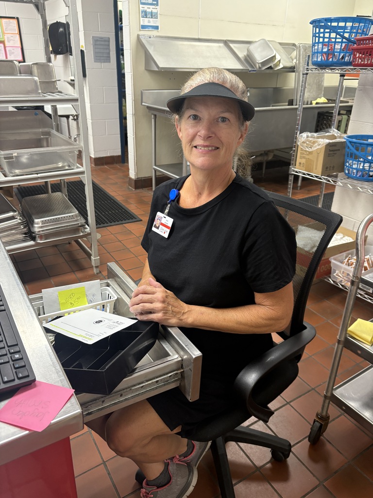 A woman sits at a desk in a kitchen with a black visor hat, black top, and gray shoes.