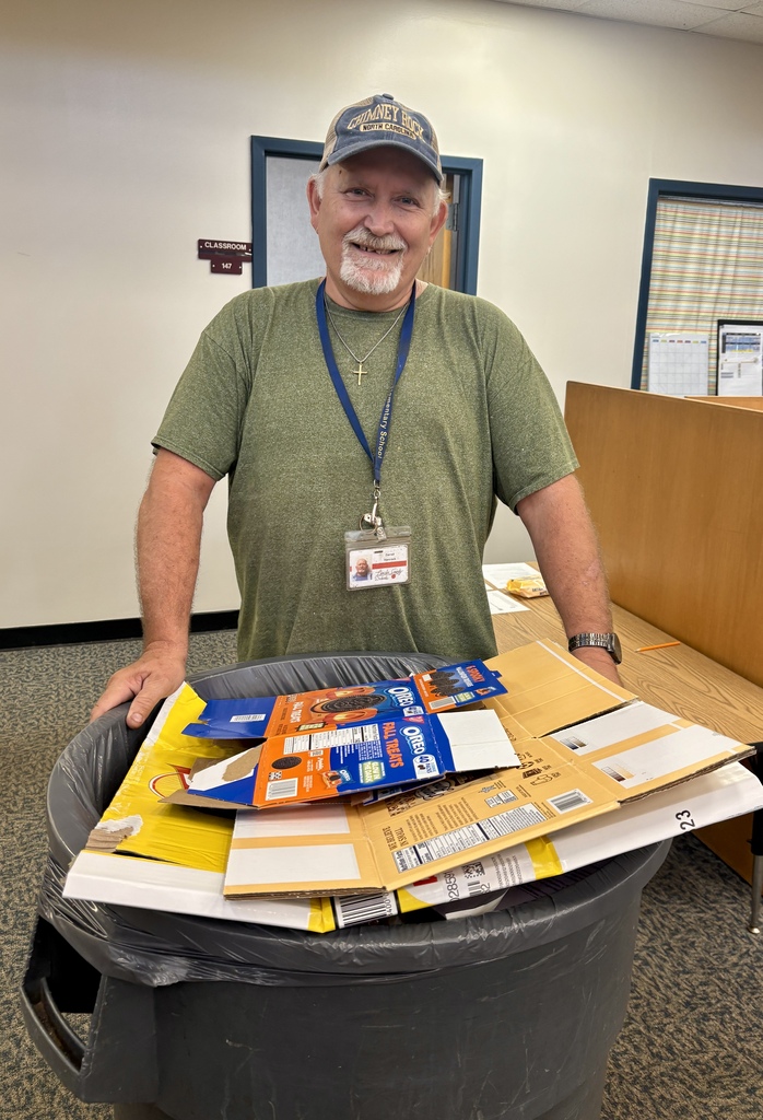 An older man in a green shirt stands in a room with a trash bin full of mail.