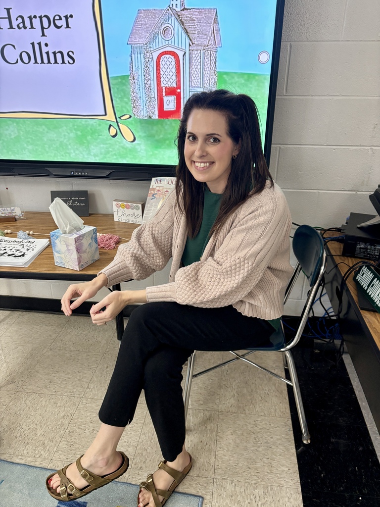 Woman sits on chair in front of classroom screen showing cartoon house. She wears a light sweater, dark pants, and sandals.