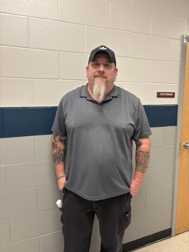 A man stands against a white wall with blue trim, wearing a gray shirt, black hat, and pants.