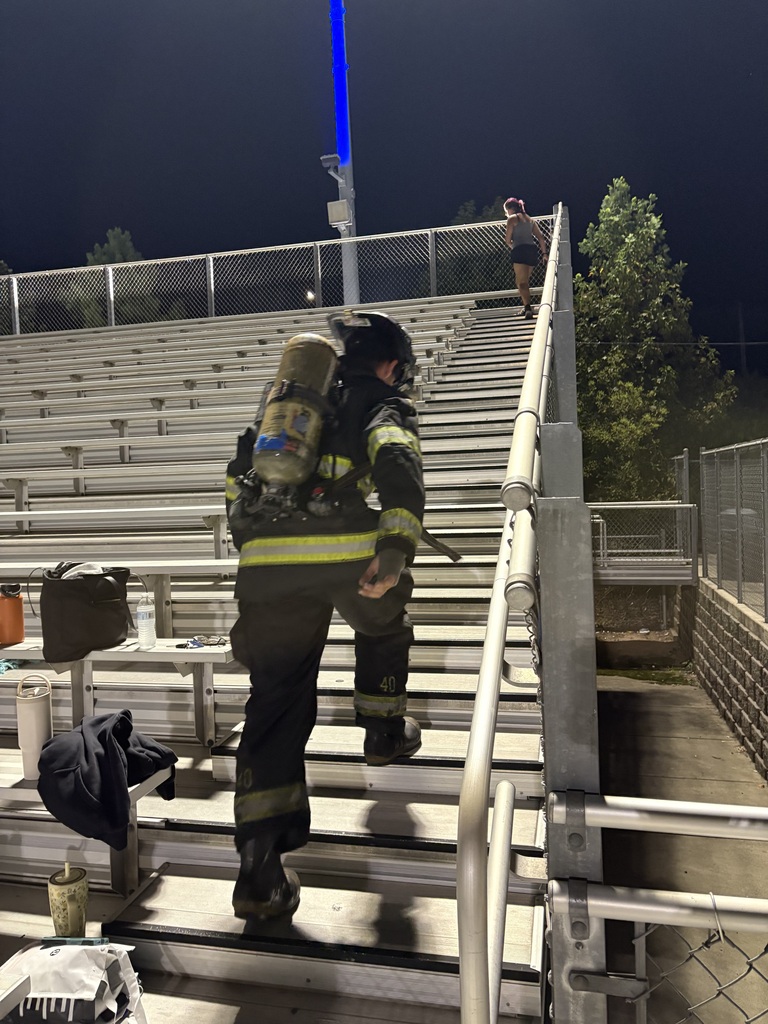 A firefighter climbs stadium steps at night, wearing a helmet and a fireproof suit. An individual walks ahead.
