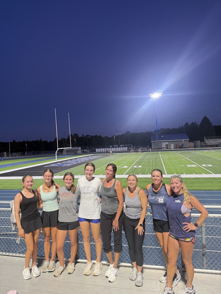 A group of women stands in front of a football field at dusk. A stadium light is on.