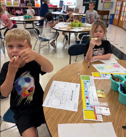 Children in a classroom eat apples. A boy is seated at a table with his fingers in his mouth.