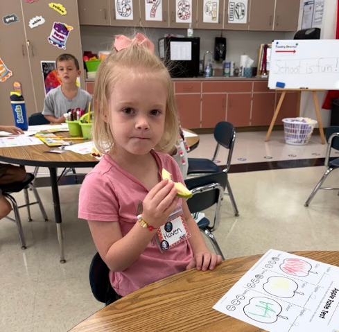 A young girl in a pink shirt sits at a table, eating an apple slice. Behind her, a classroom has tables, chairs, and a whiteboard.